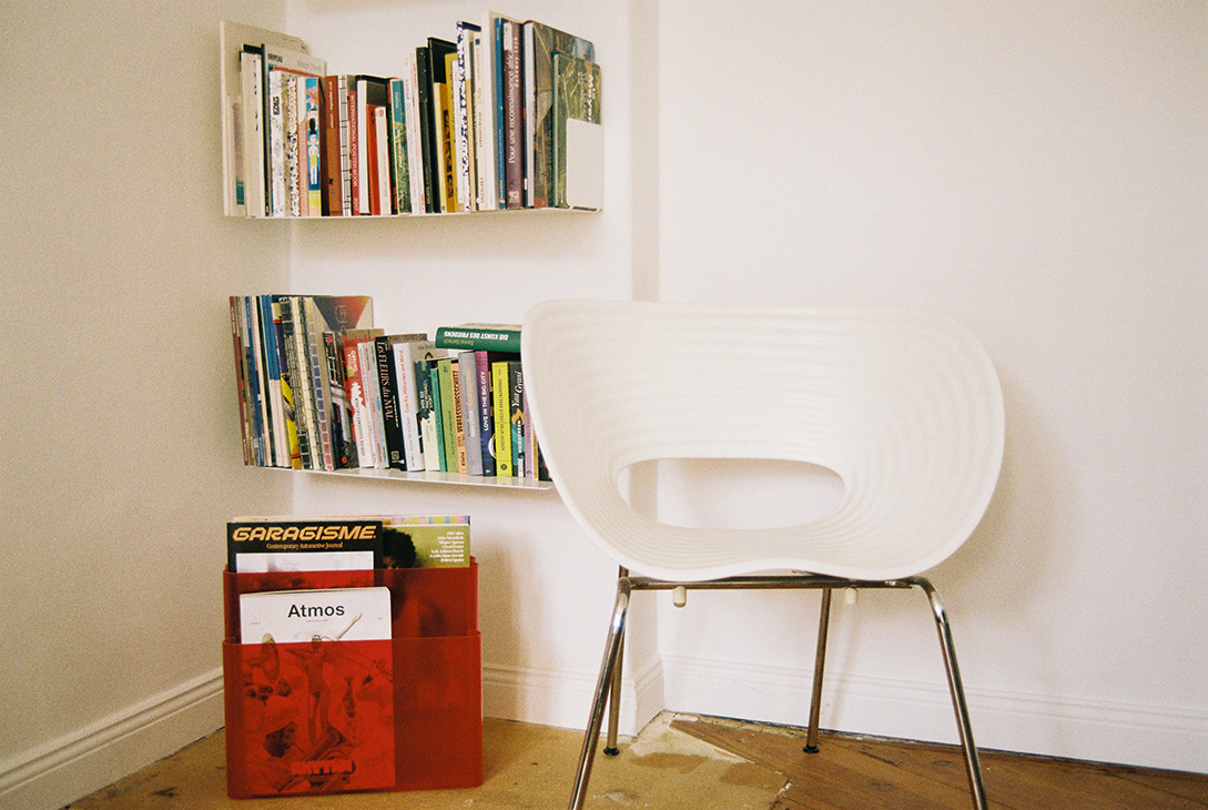 A photo of a corner of a room. Two bookshelves filled with books hang on the wall. On the floor is a red magazine rack containing various magazines and a white chair.